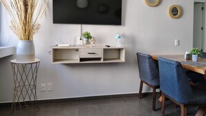 Un soggiorno moderno con una televisione a schermo piatto, una piccola mensola con oggetti decorativi, un tavolo da pranzo con sedie e un'alta vaso con piante essiccate.