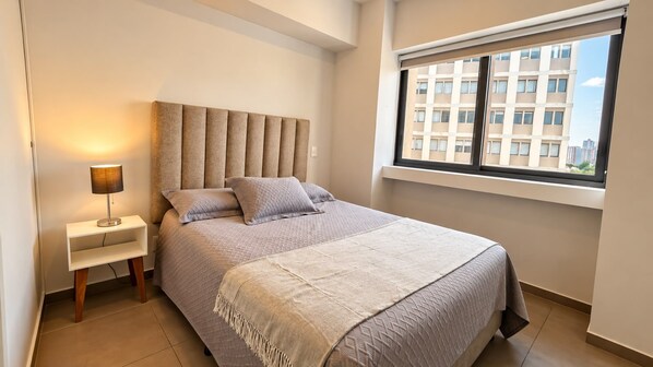 A bedroom with a bed, a bedside lamp, a window, and a view of buildings outside. - App. Lafayette 18B: 2 bedrooms, 1 bathroom, 1 parking space (Guadalajara)