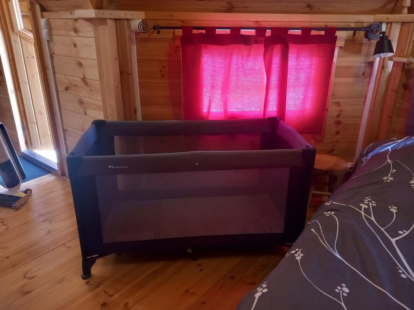A wooden cabin room with a bed, a small table, and a window with a red curtain.
