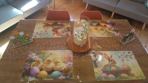 Een gezellige kamer met twee banken, een houten eettafel met tafelonderleggers met paasthema, en een klein bijzettafeltje met decoratieve objecten.