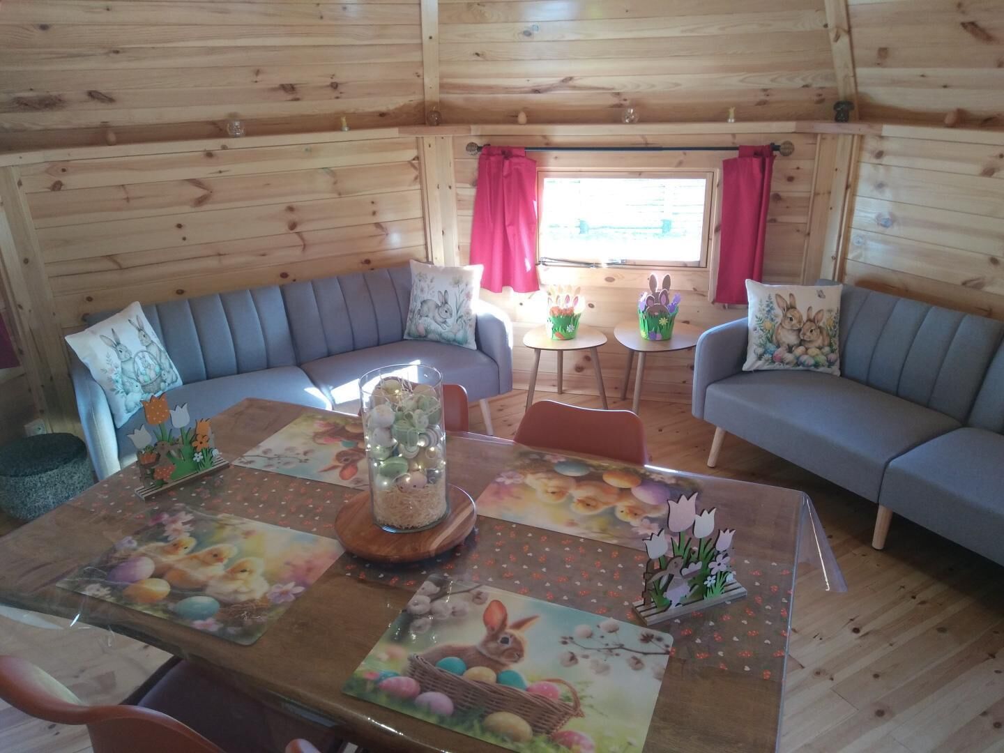 A cozy cabin interior with a wooden interior, a sofa, a dining table with Easter-themed placemats, and a window with curtains.