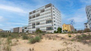 Een meerlaags hotel met balkons, gelegen nabij een strand met duinen en schaarse begroeiing.