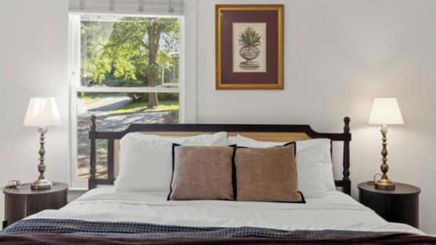 A neatly made bed with white and brown bedding, a framed picture on the wall, and two bedside lamps. - New listing! The Terra Cotta Bungalow with Firepit! (Athens)