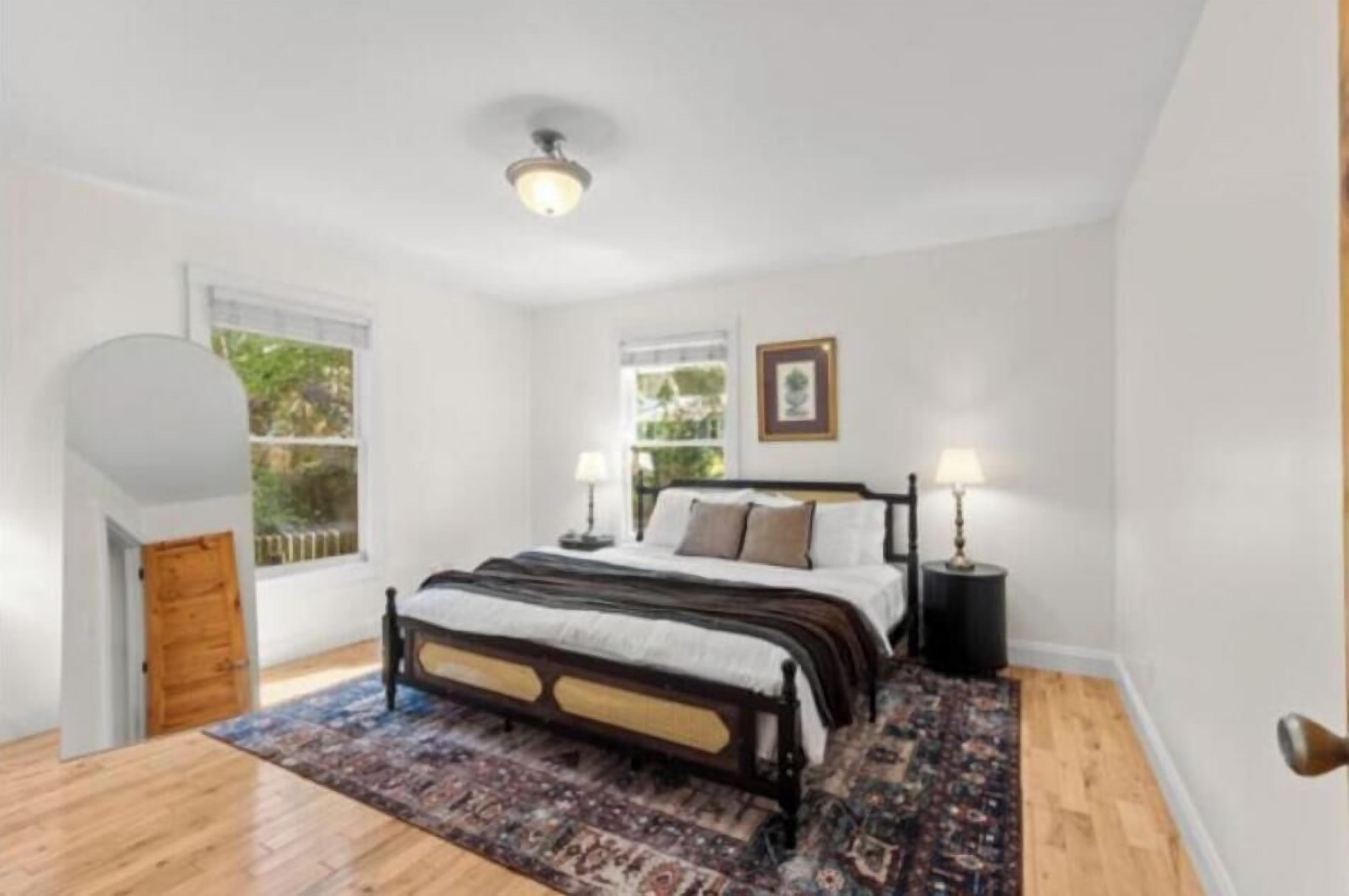 A bedroom with a large bed, two bedside tables with lamps, a window with a view of greenery, and a door leading to another room.