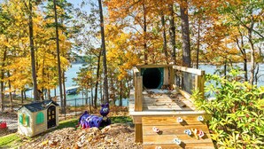 A wooden deck with a tunnel entrance, a small playhouse, and a purple toy figure. - Hartwell Haus with Dock & Playground (Fair Play)