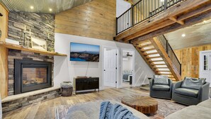 A spacious living room with a fireplace, a stone wall, a wooden ceiling, and a staircase.