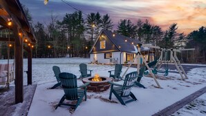 A cozy cabin with a fire pit and outdoor seating, surrounded by snow and trees.