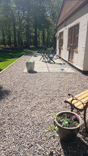Un espace patio avec un sol en gravier, un banc en bois, une plante en pot et un barbecue à l’arrière-plan.