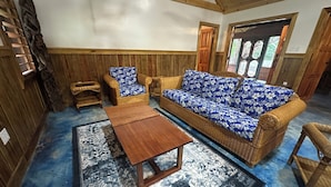 Una habitación con techo de madera, un sofá azul con estampado floral, dos sillas de mimbre, una mesa de centro de madera y una alfombra estampada.