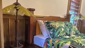 Un dormitorio con cabecera de madera, cama con colcha de motivos tropicales, lámpara de noche y un cubo de basura.