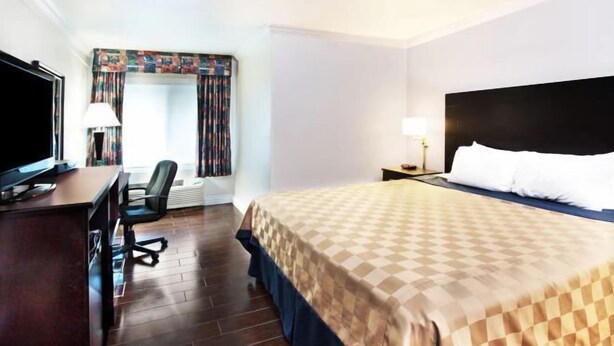 A hotel room with a bed, a desk with a TV, a chair, a nightstand, and a window with curtains. - Travelodge by Wyndham Sunset-Huntington Beach Ocean Front (Sunset Beach)