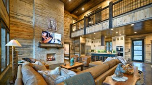 A spacious living area with a large leather sofa, a fireplace, and a television mounted on the wall. - Wild & Free | Luxury 7BR Cabin w/ Heated Pool, Pickleball Court, Game Loft & Multiple King Suites (Broken Bow)