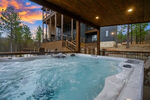 A hot tub with wooden deck and stairs leading to a building with a balcony. - Wild & Free | Luxury 7BR Cabin w/ Heated Pool, Pickleball Court, Game Loft & Multiple King Suites (Broken Bow)