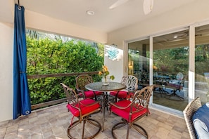 Une terrasse avec une table ronde, des chaises à coussins rouges et une vue sur la verdure, visible à travers une porte vitrée.