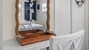 Une pièce avec un plateau en bois posé sur une console blanche, une chaise blanche et un grand miroir qui reflète un lit et une fenêtre.
