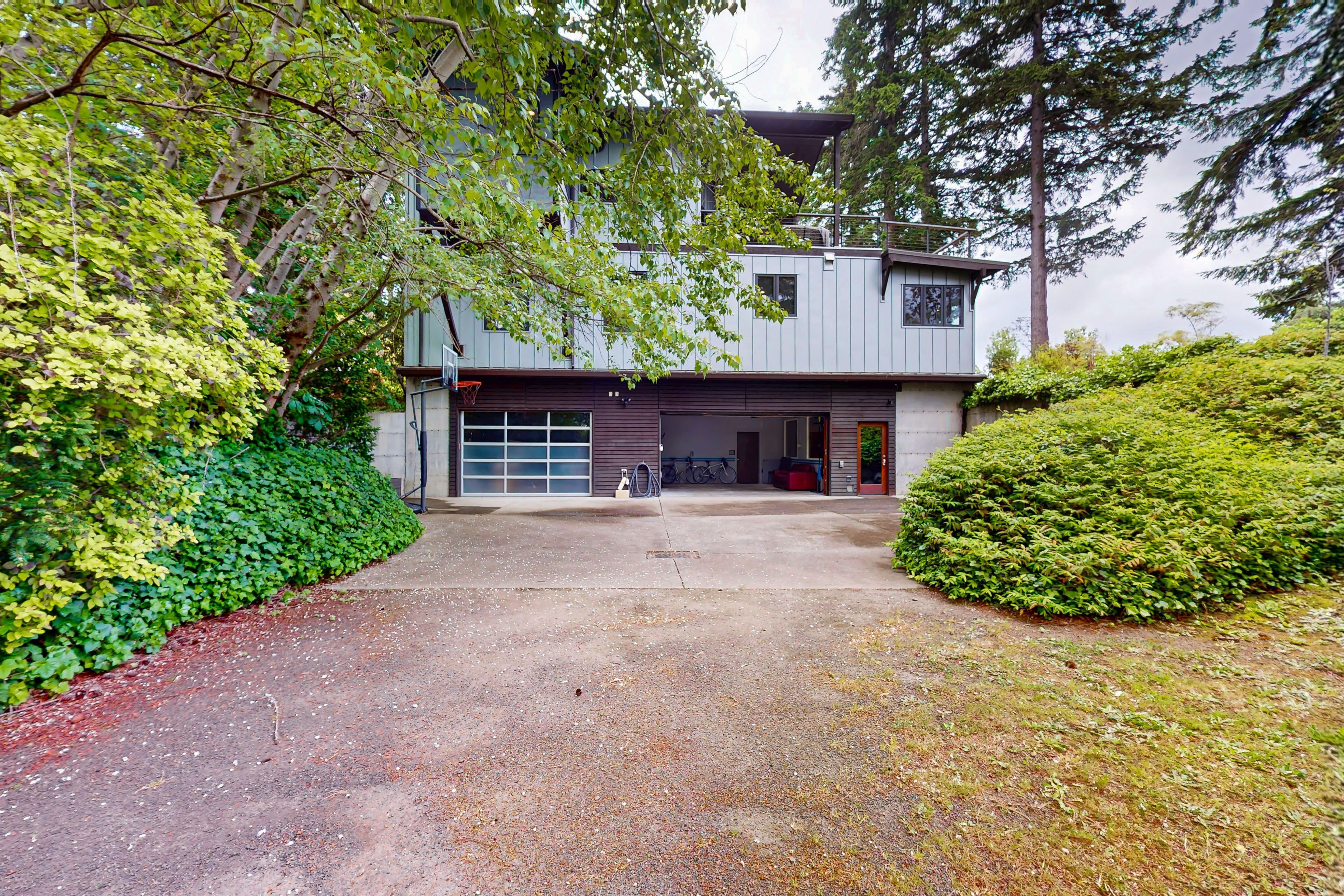 Une maison moderne à deux étages avec un garage, entourée d’une végétation luxuriante et d’arbres.