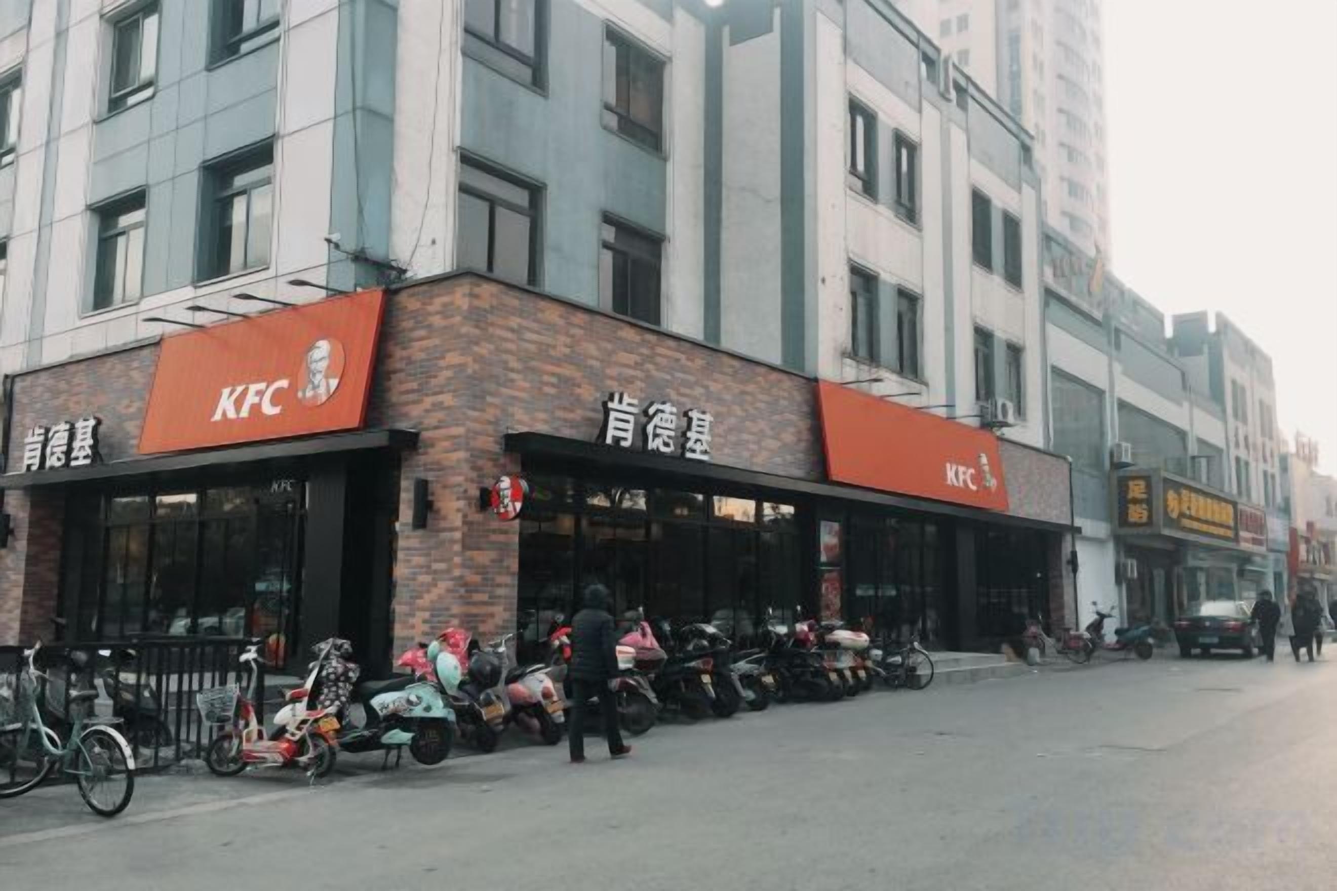 Restaurante KFC com mesas ao ar livre, bicicletas estacionadas na frente e um edifício com várias janelas ao fundo.
