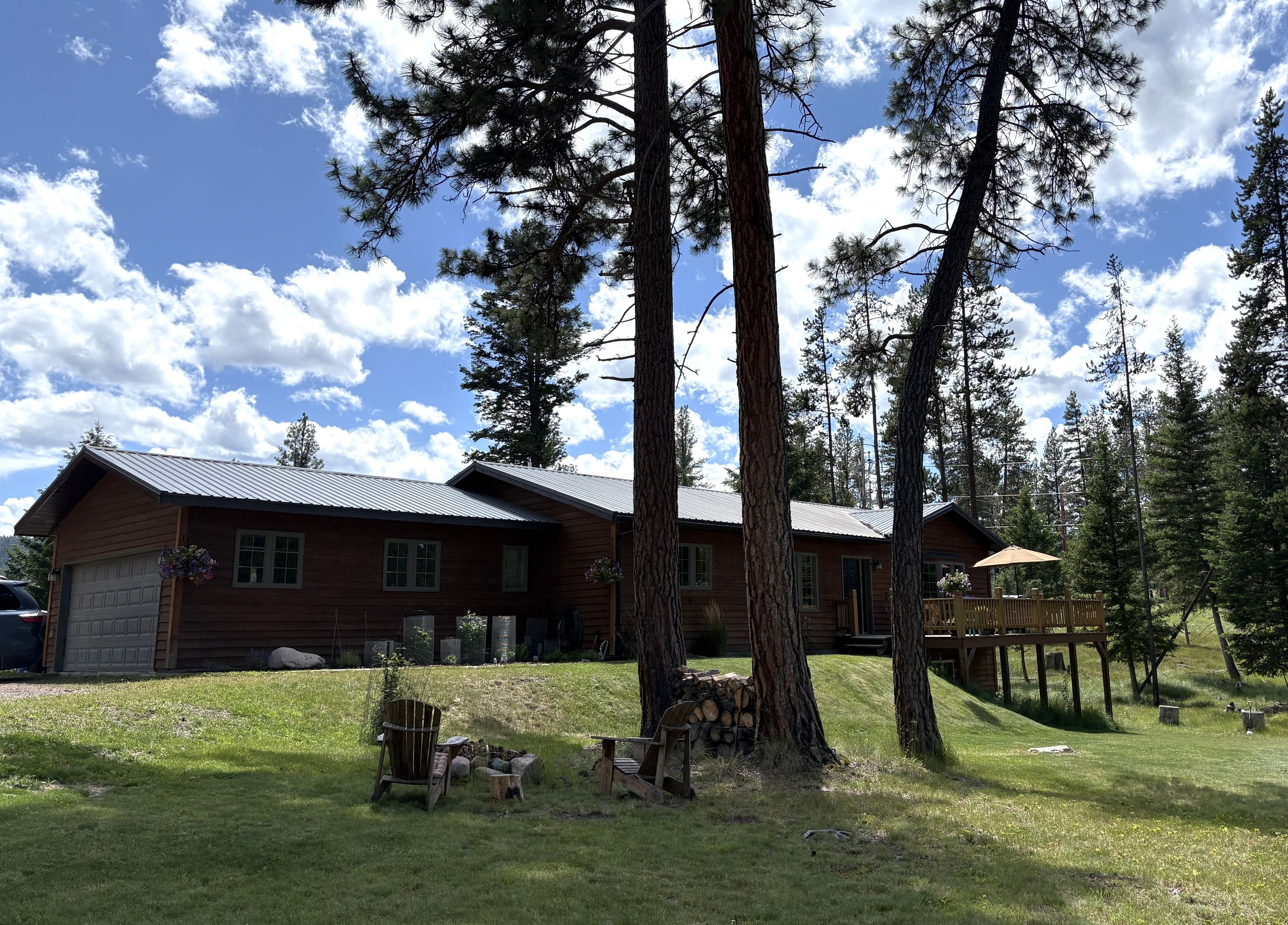 A two-story log cabin with a garage, surrounded by tall pine trees and a clear sky.