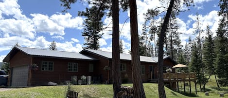 A two-story log cabin with a garage, surrounded by tall pine trees and a clear sky.