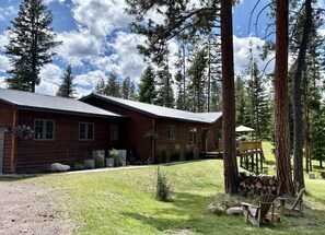 A wooden cabin with a metal roof, surrounded by a grassy area and tall pine trees. - Charming 3-bedroom house in Seeley Lake for a relaxing stay (Seeley Lake)