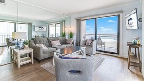 Ein modernes Wohnzimmer mit großem Fenster zum Meer, einem grauen Sofa, einem Couchtisch und einem kleinen Beistelltisch.