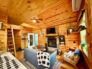 Ein Blockhaus-Interieur mit Kamin, einer Couch, einer Leiter und einem Fernseher.