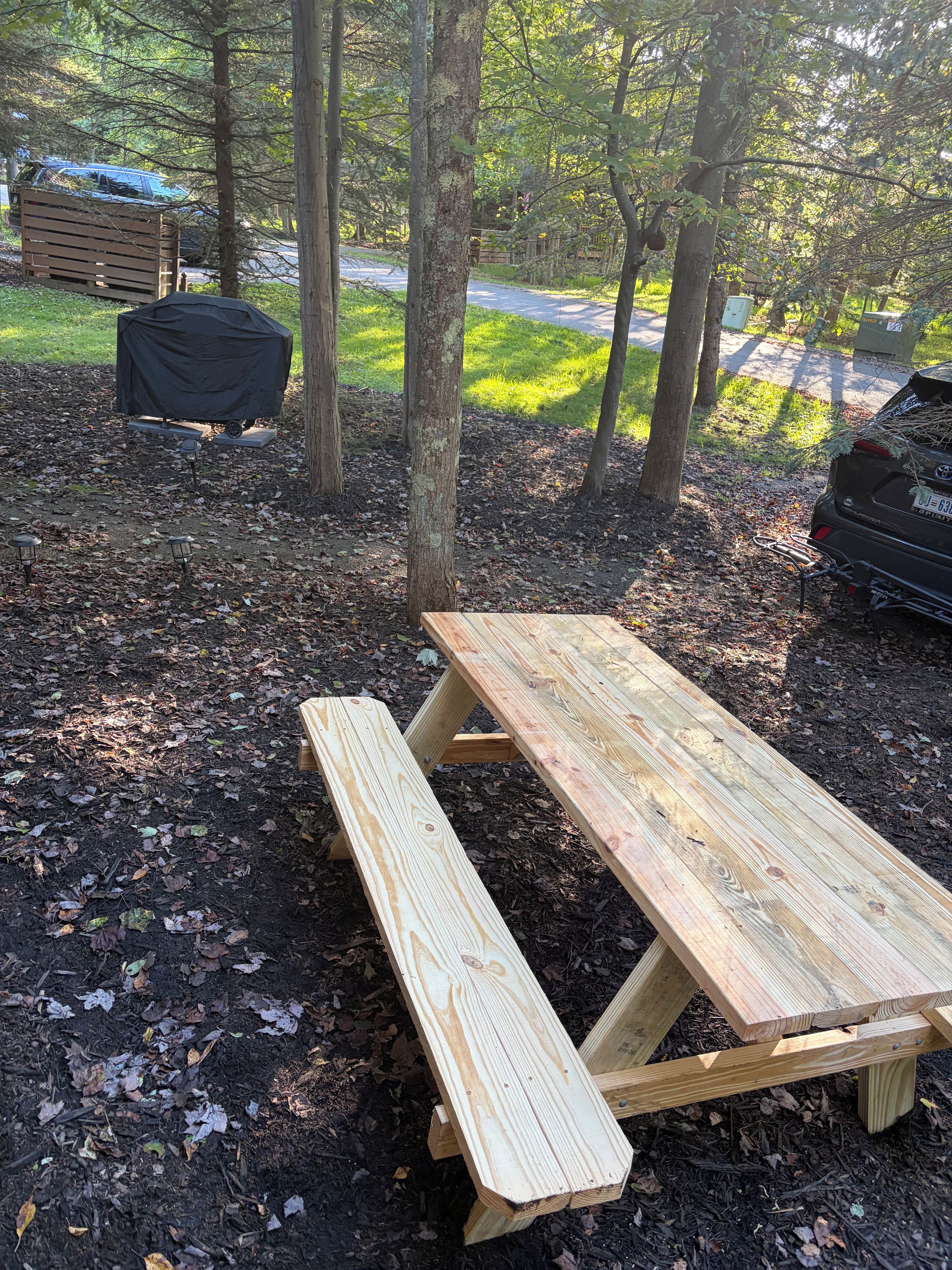 A picnic table with benches in a wooded area with trees and a road in the background.