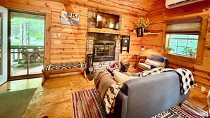 A wooden cabin interior with a stone fireplace, a sofa, a bench, and a large window with plants.