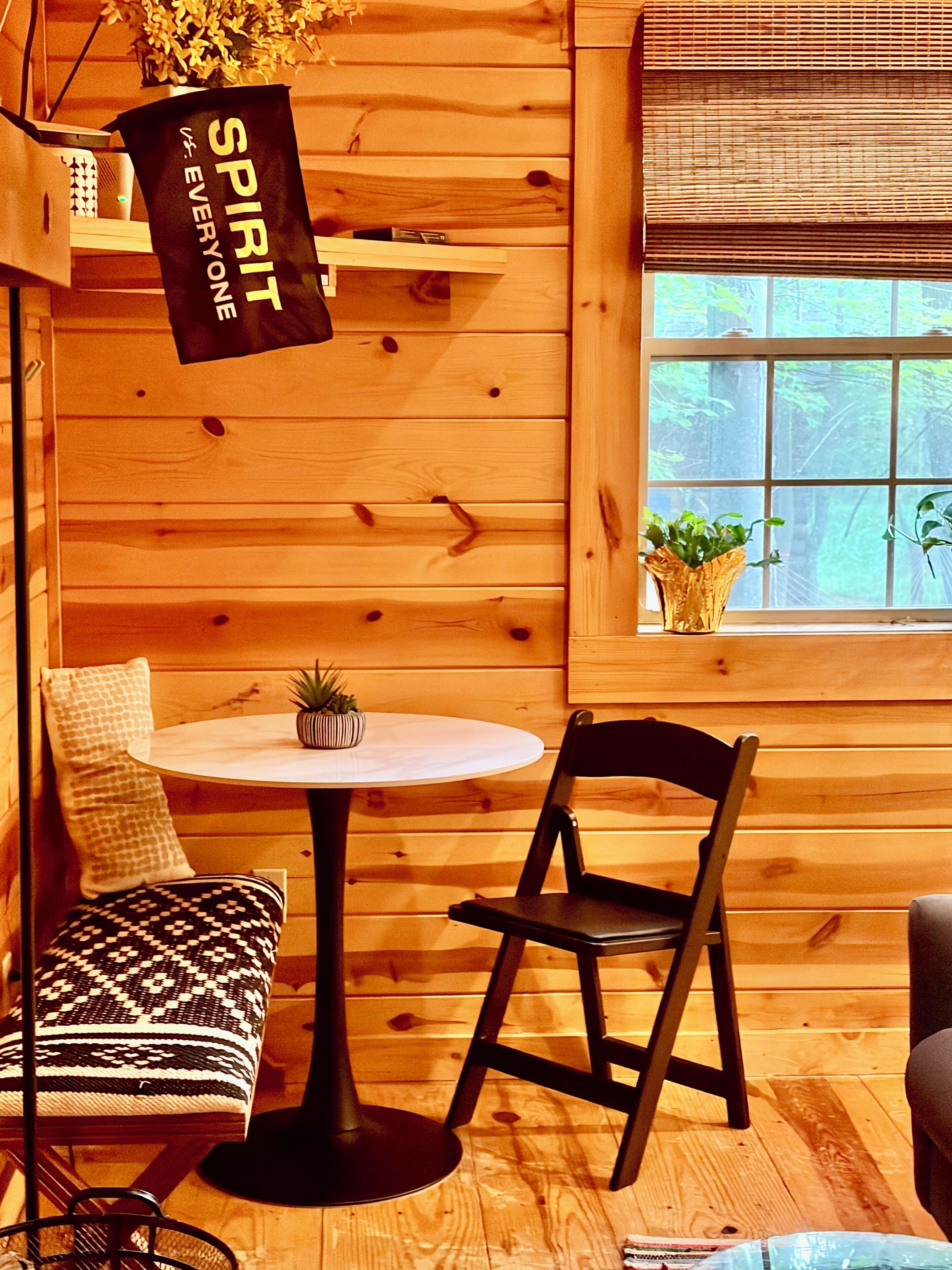 A wooden interior with a round table, two chairs, a small plant, and a banner.