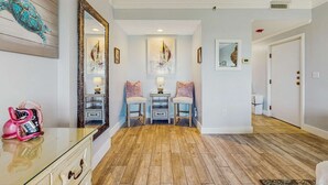 A hallway with wooden flooring, a glass table with a pink kettle, two chairs with lamps, and artwork on the walls. - Gulf view studio for 2 with balcony (Miramar Beach)
