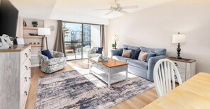 A modern living room with a sofa, coffee table, and a dining table. There is a large window with a view of buildings outside. - Bluewater-Modern 1BR Oasis with Pool & Easy Beach Access (Ocean City)