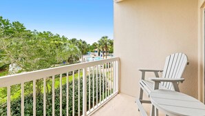 A balcony with a white railing, two Adirondack chairs, and a view of a swimming pool and greenery. - 1BR condo steps to beach, pools, hot tub (Miramar Beach)