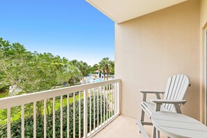 A balcony with a white railing, two Adirondack chairs, and a view of a swimming pool and greenery. - 1BR condo steps to beach, pools, hot tub (Miramar Beach)