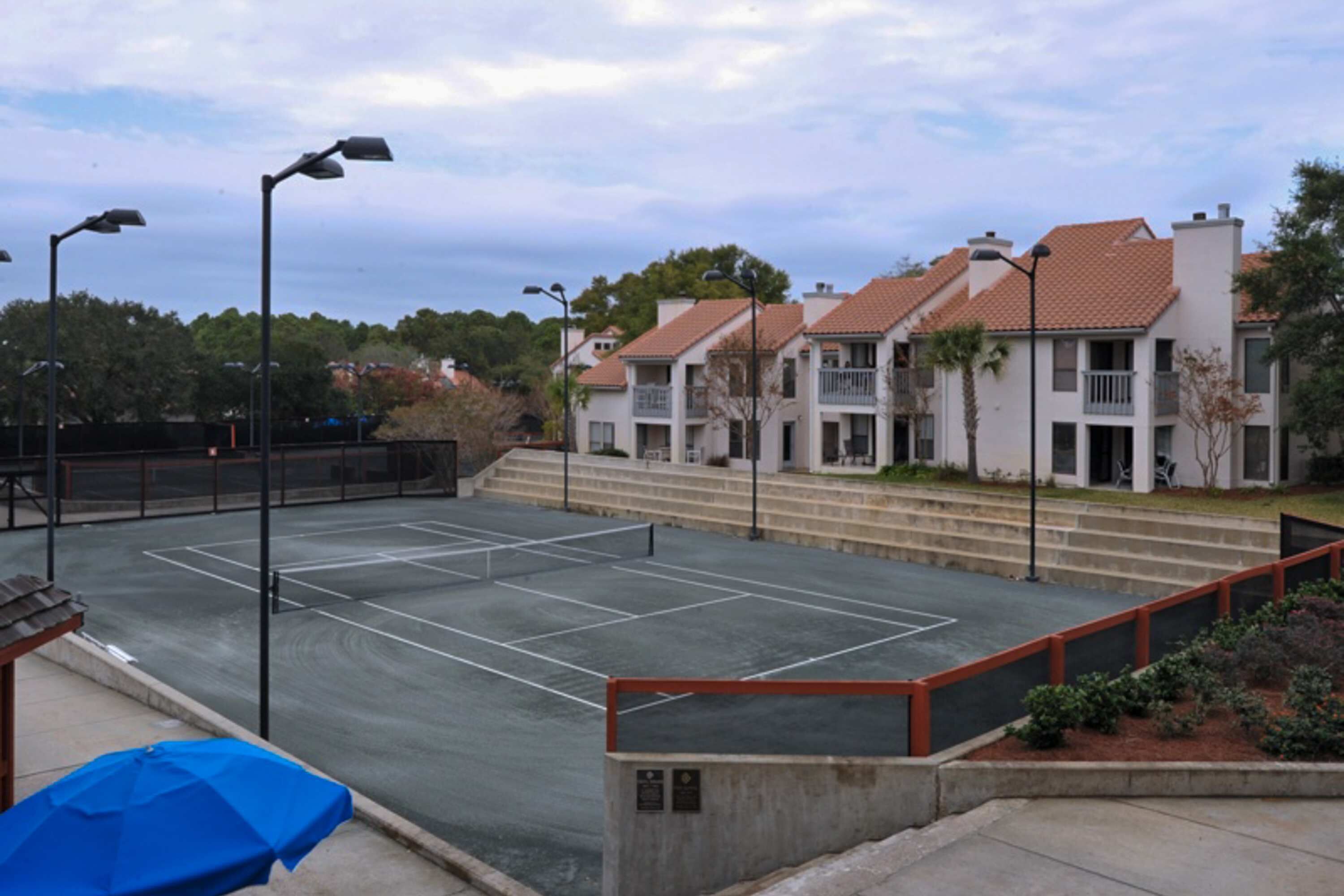 A tennis court with a net, surrounded by a low fence and steps leading to a building with balconies.