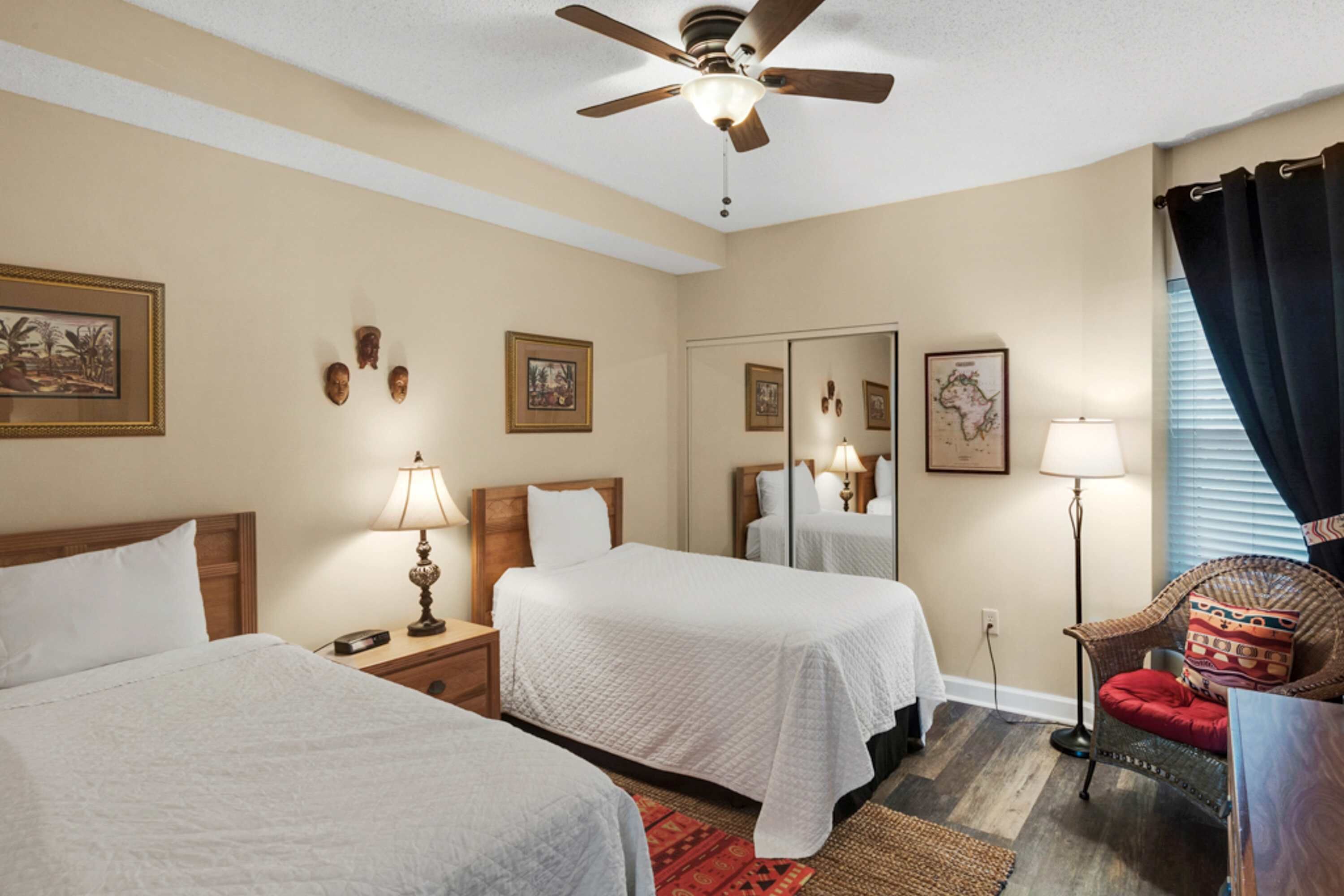 A hotel room with two beds, a ceiling fan, a mirror, a chair, and a lamp.