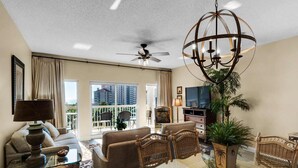 A spacious living room with a ceiling fan, a large chandelier, and a view of the city through large windows. - Luxury condo w/private balcony & resort access (Miramar Beach)