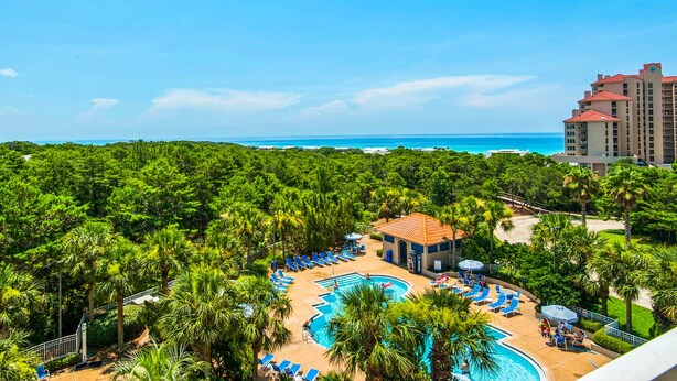 Un resort con piscina rodeada de palmeras, vista a la playa y un edificio grande al fondo.