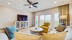 Ein geräumiges Wohnzimmer mit Deckenventilator, einem runden Couchtisch, Sofas, einem Fernseher und Blick ins Freie.