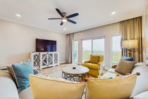 Ein geräumiges Wohnzimmer mit Deckenventilator, einem runden Couchtisch, Sofas, einem Fernseher und Blick ins Freie.