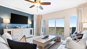 A modern living room with a flat-screen TV, a ceiling fan, a sofa, a coffee table, and a view of the outdoors. - Steps to beach, ocean views, pool access (Miramar Beach)
