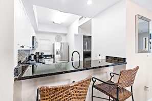 A modern kitchen with a glass island, stainless steel appliances, and wicker chairs. - Steps to beach, ocean views, pool access (Miramar Beach)