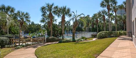 A paved pathway, palm trees, a pool, and a building with a fence.