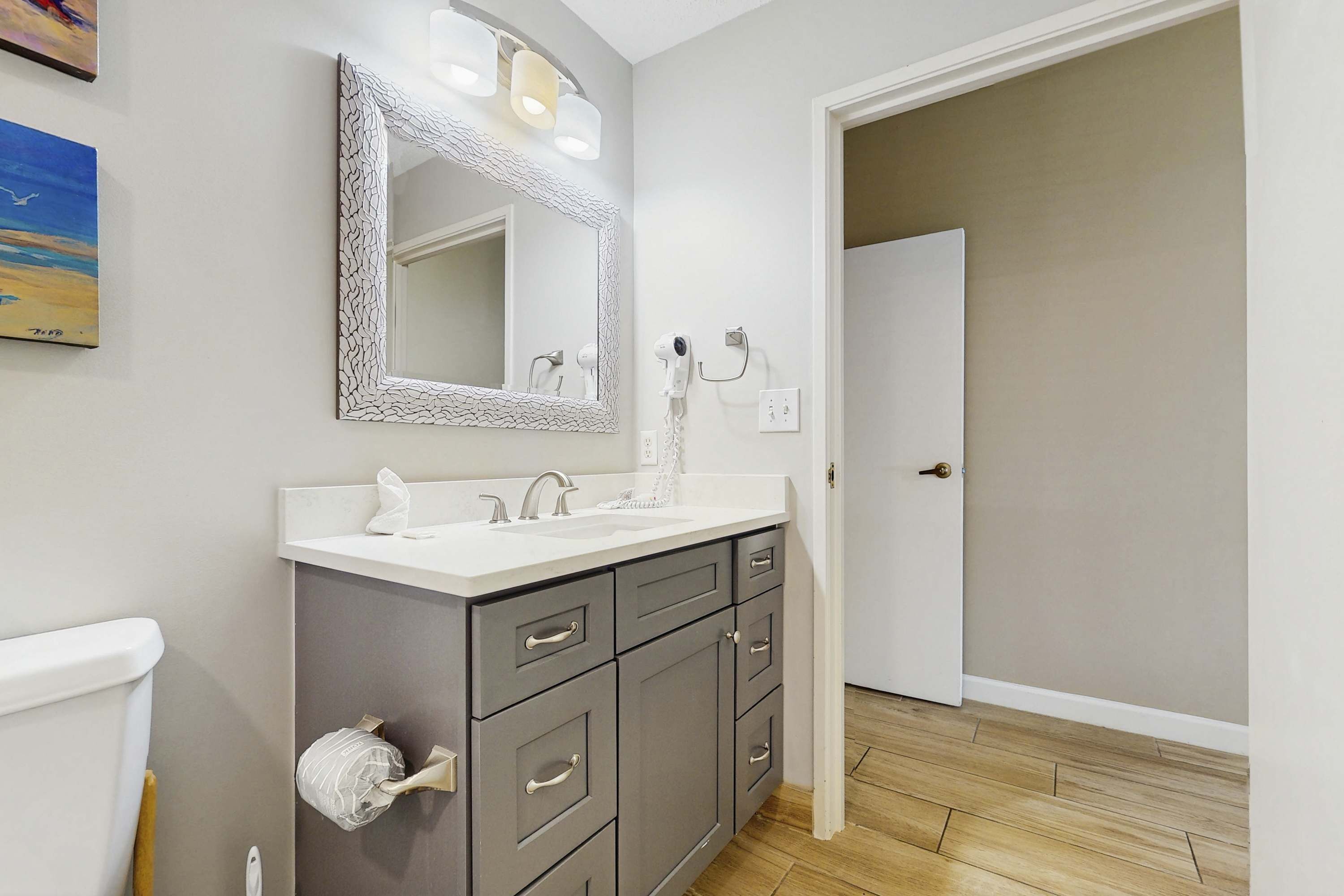 Une salle de bain avec un lavabo blanc, un miroir doté d’un cadre texturé, un sèche-cheveux et des toilettes.