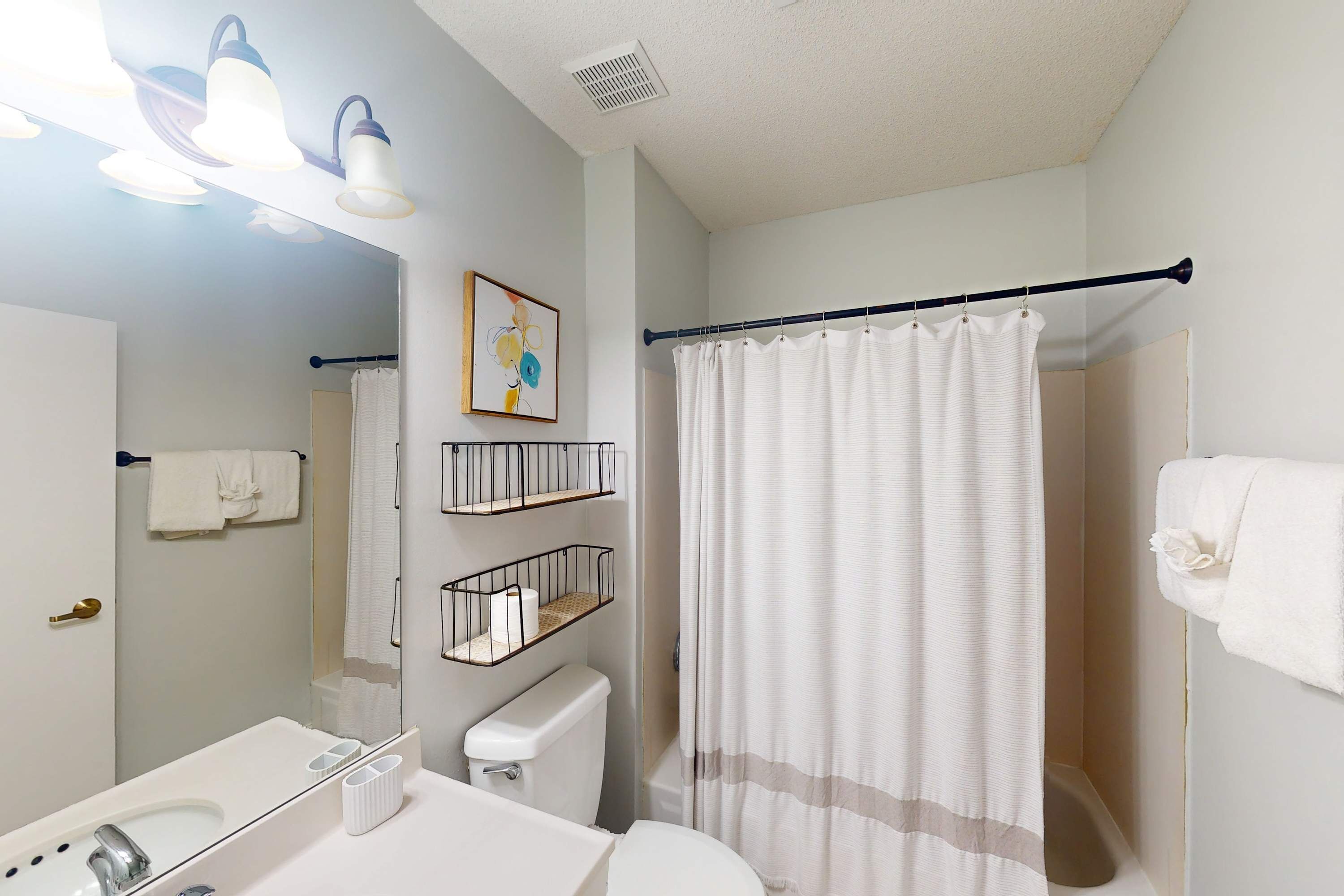 A bathroom with a toilet, sink, mirror, shower curtain, towel rack, and wall-mounted shelves.