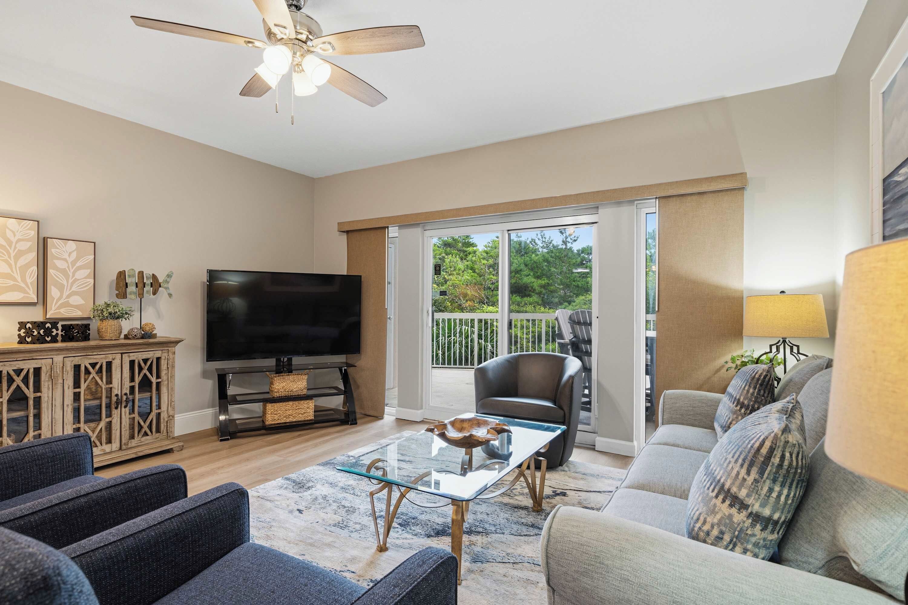 A spacious living room with a flat-screen TV, comfortable seating, a ceiling fan, and a view through a sliding glass door.