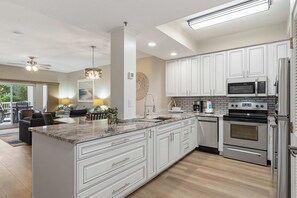 A modern kitchen with white cabinetry, a central island, stainless steel appliances, and a dining area with a view. - Gulf-Front Condo with Pool Fitness and Hot Tub (Miramar Beach)