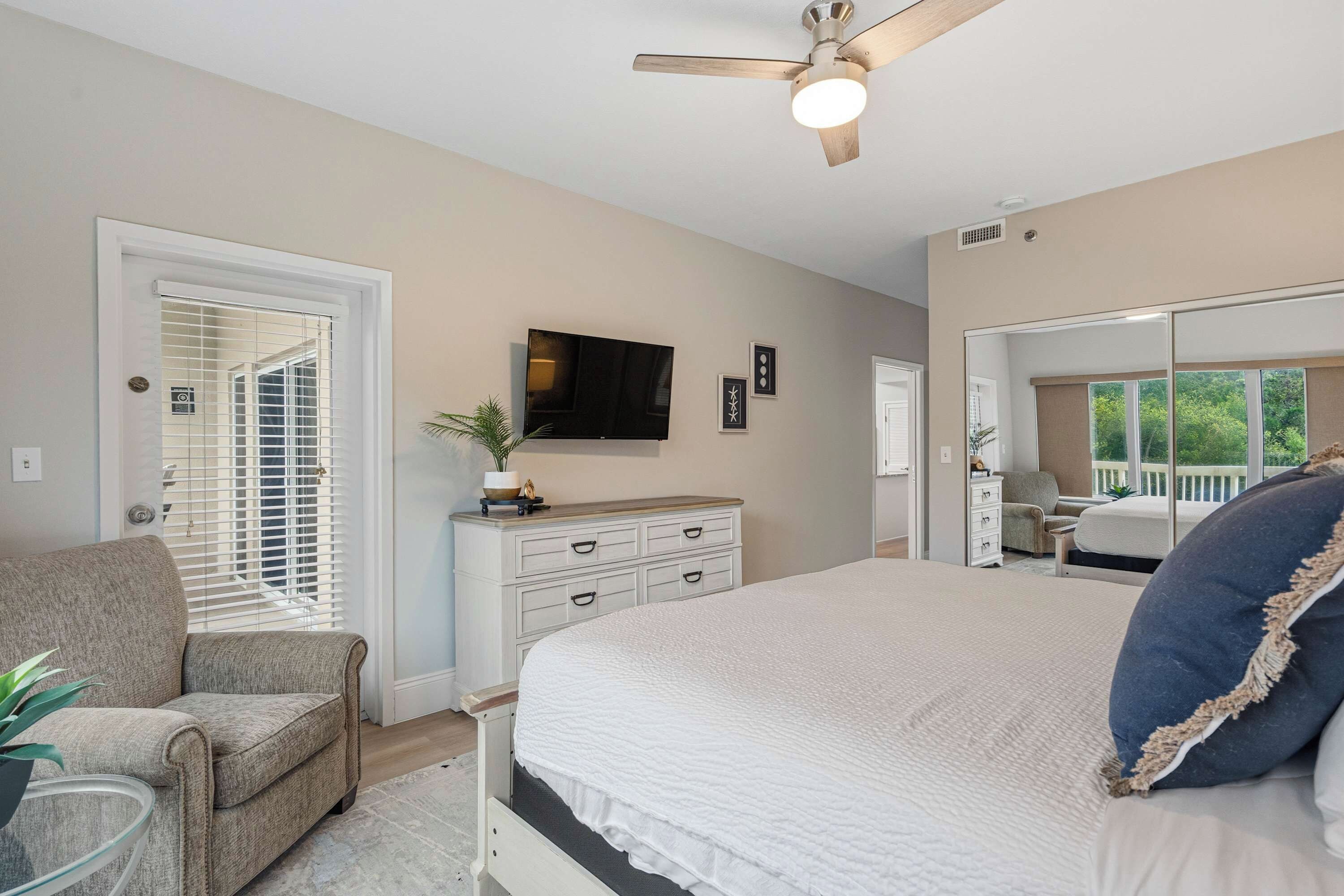 A bedroom with a large bed, a ceiling fan, a television, and a view of the outdoors through a window.