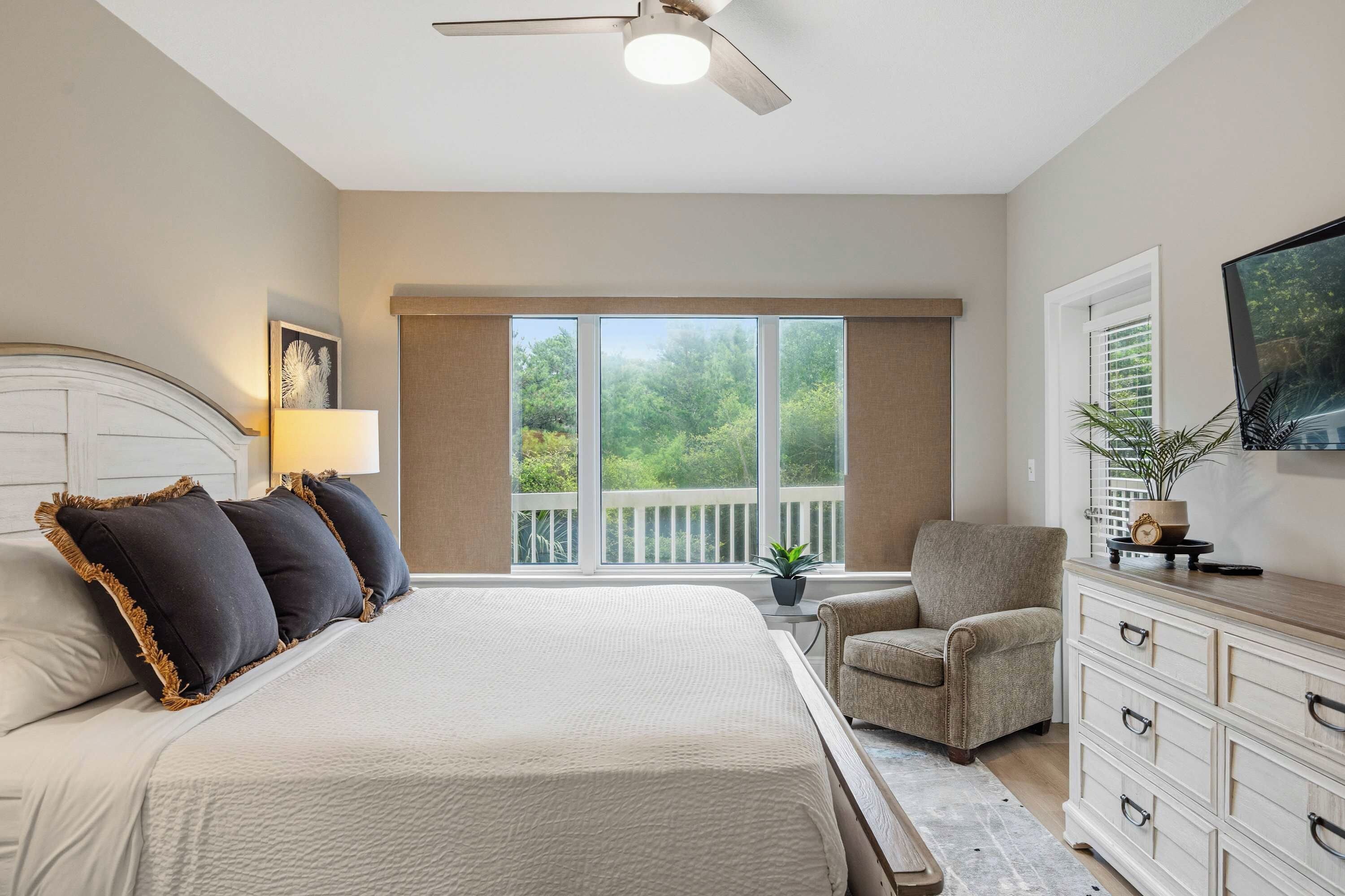 A bedroom with a large bed, a chair, a dresser, a television, and a balcony with a view of trees.