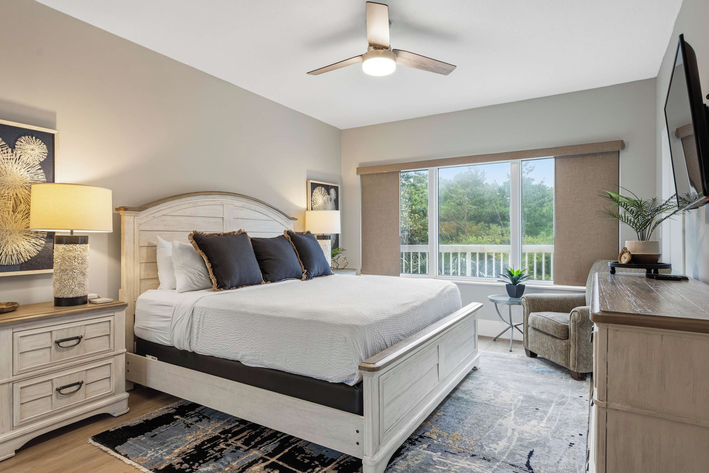 A bedroom with a bed, bedside tables, a ceiling fan, a window with a view, and a small table with a plant.