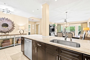 A modern kitchen with a built-in dishwasher, a large island with a sink, and a dining area with chairs and a table. - Exclusive 2BR: Steps to Beach, Pools, Hot Tub (Miramar Beach)